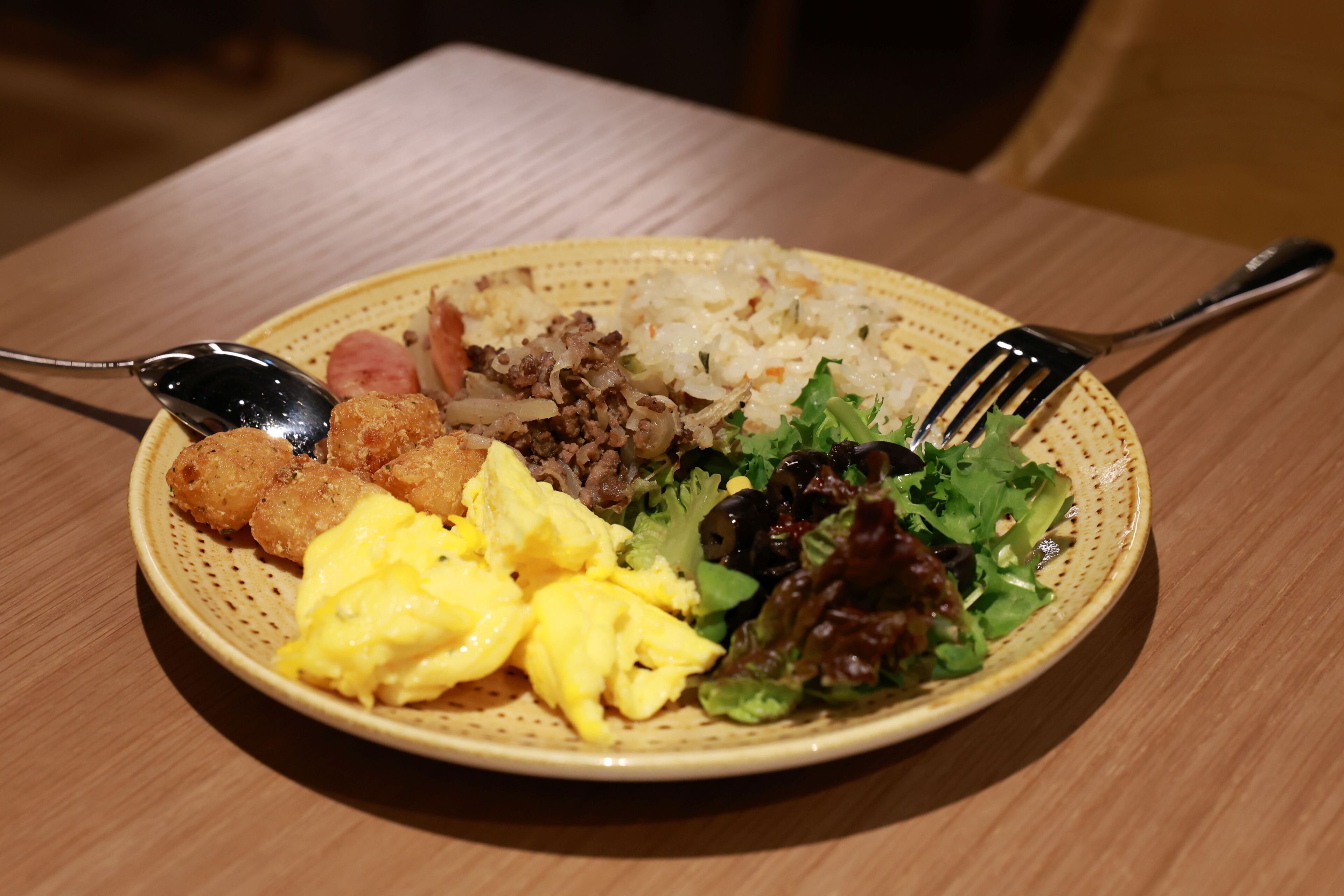 Ein Teller mit Rührei, Hackfleisch, Reis, Kartoffelwürfeln und einem gemischten Salat auf einem Holztisch.