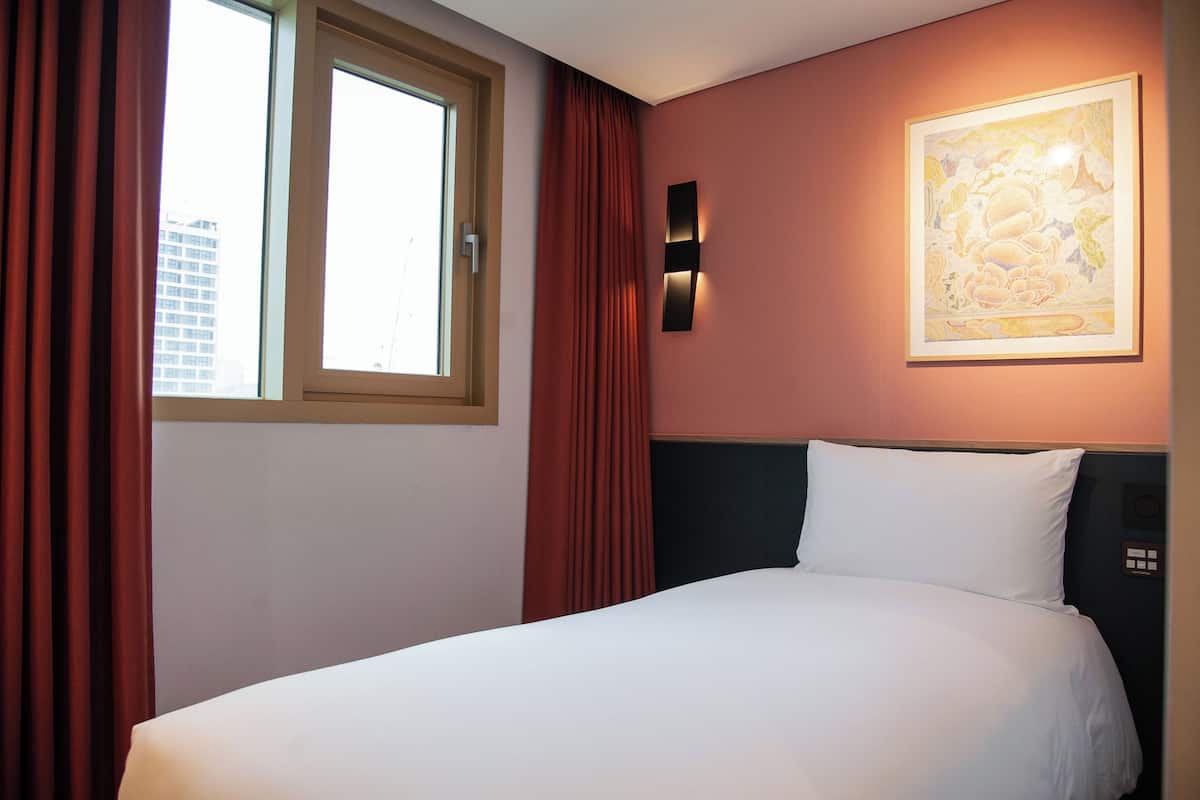 A hotel room with a bed, a window with curtains, a wall-mounted light, and a framed artwork.