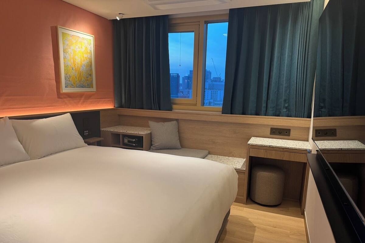 A hotel room with a large bed, a desk, a chair, and a small trash bin.