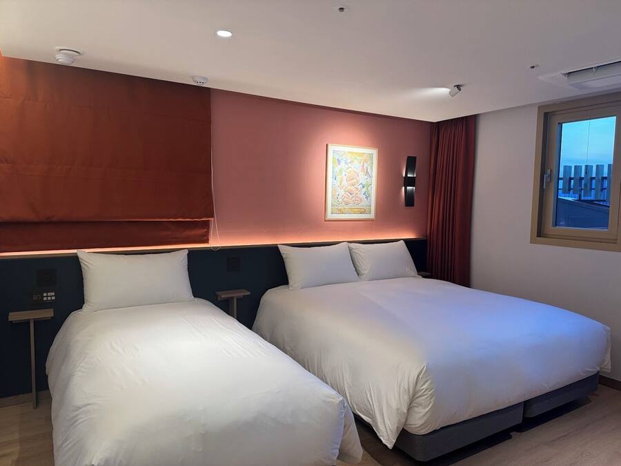 A hotel room with two beds, a painting on the wall, a window with a view, and a control panel on the wall.