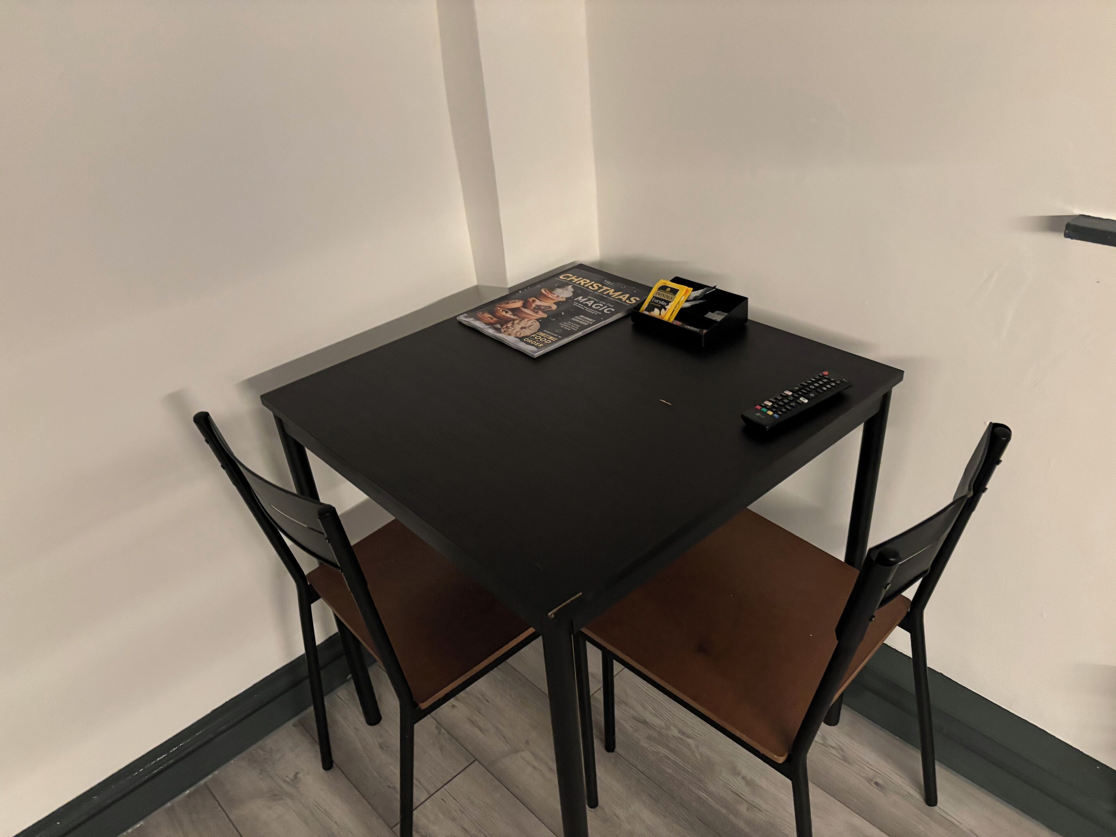 A small, dark wooden table with a magazine, a remote control, and a small black box. Two black chairs are positioned around the table.