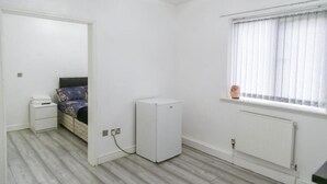A bedroom with a bed, a small white refrigerator, a nightstand, a window with blinds, and a radiator. - Hill's View Apartments (Manchester)