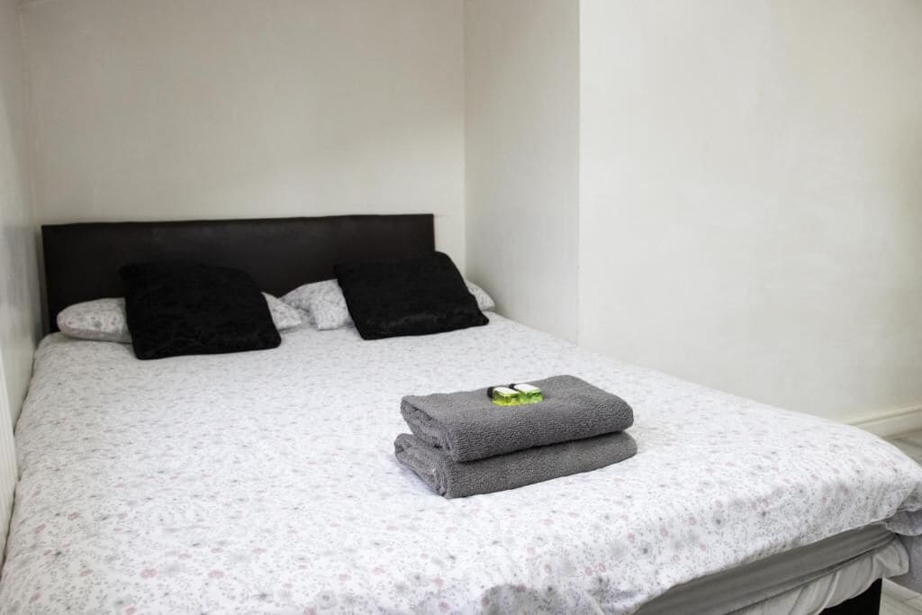 Cama bem arrumada com colcha estampada em florais, duas almofadas pretas e uma toalha cinza dobrada com um objeto verde em cima.
