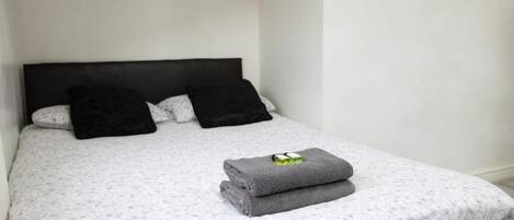 Cama bem arrumada com colcha estampada em florais, duas almofadas pretas e uma toalha cinza dobrada com um objeto verde em cima.