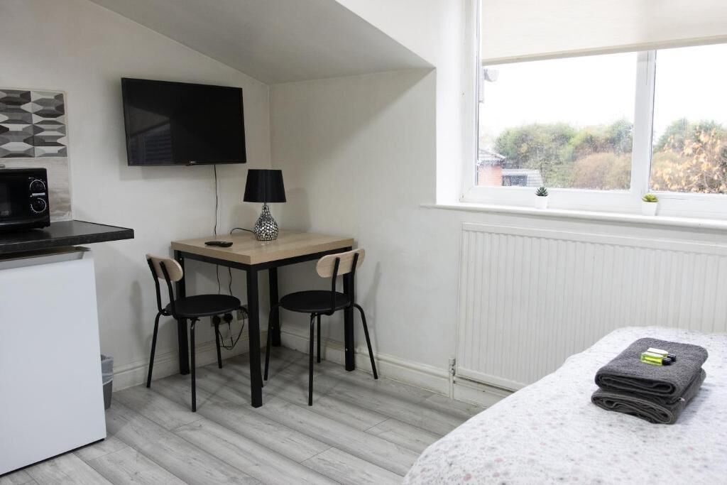 Habitación pequeña con cama, escritorio, dos sillas, televisor, lámpara y ventana con vista a árboles.