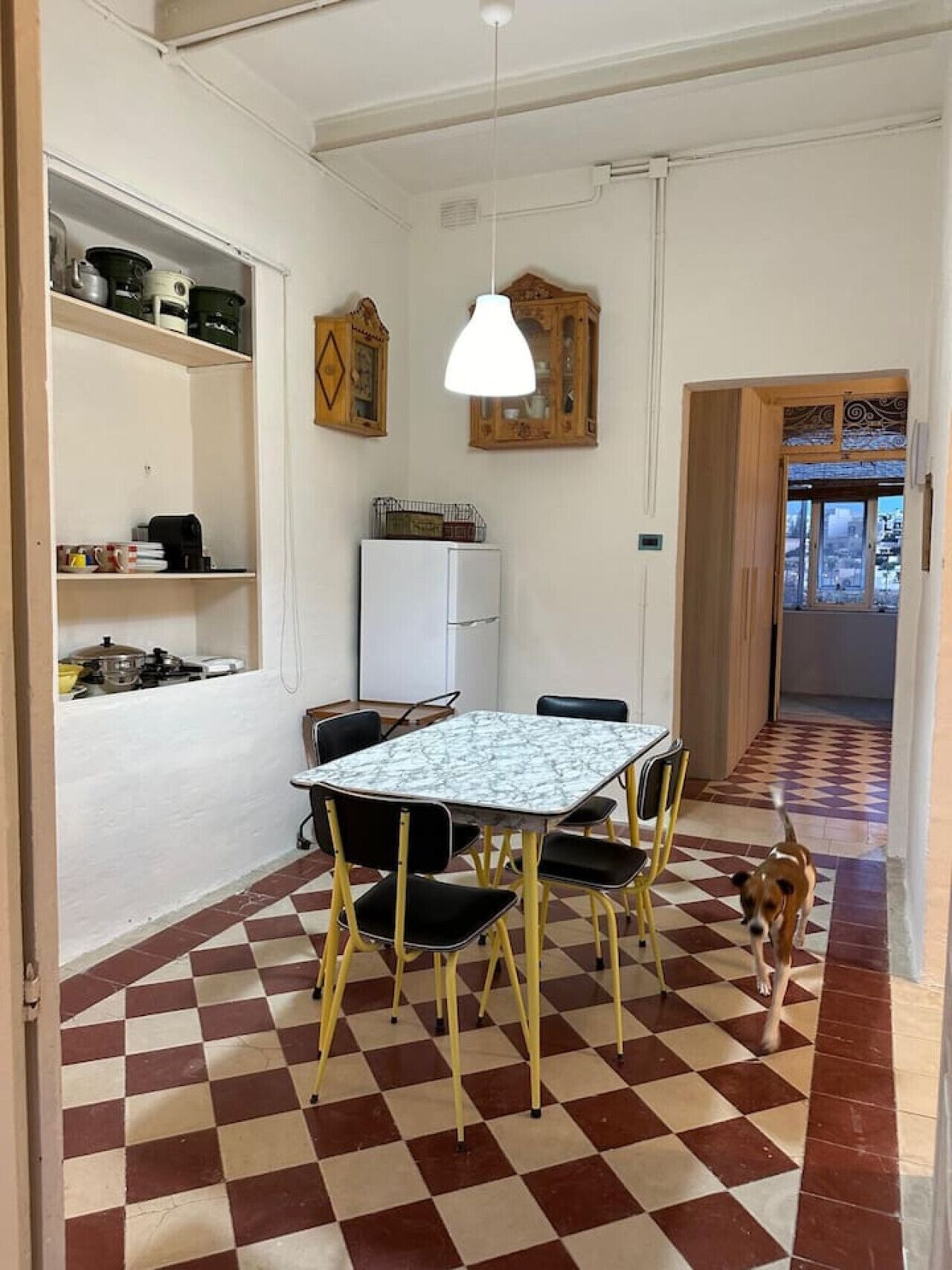 A dining area with a checkered floor, a table with chairs, a refrigerator, and a hanging light fixture.