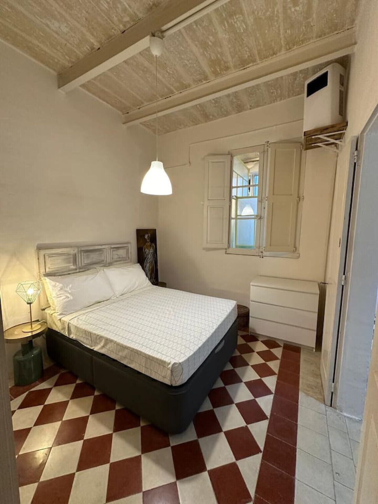 A bedroom with a checkered floor, a bed with a white mattress and dark headboard, a hanging light fixture, a window with shutters, and a white wardrobe.