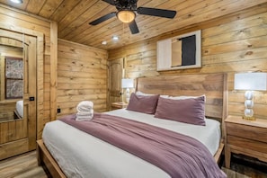 A wooden room with a bed, a ceiling fan, and a nightstand with a lamp.