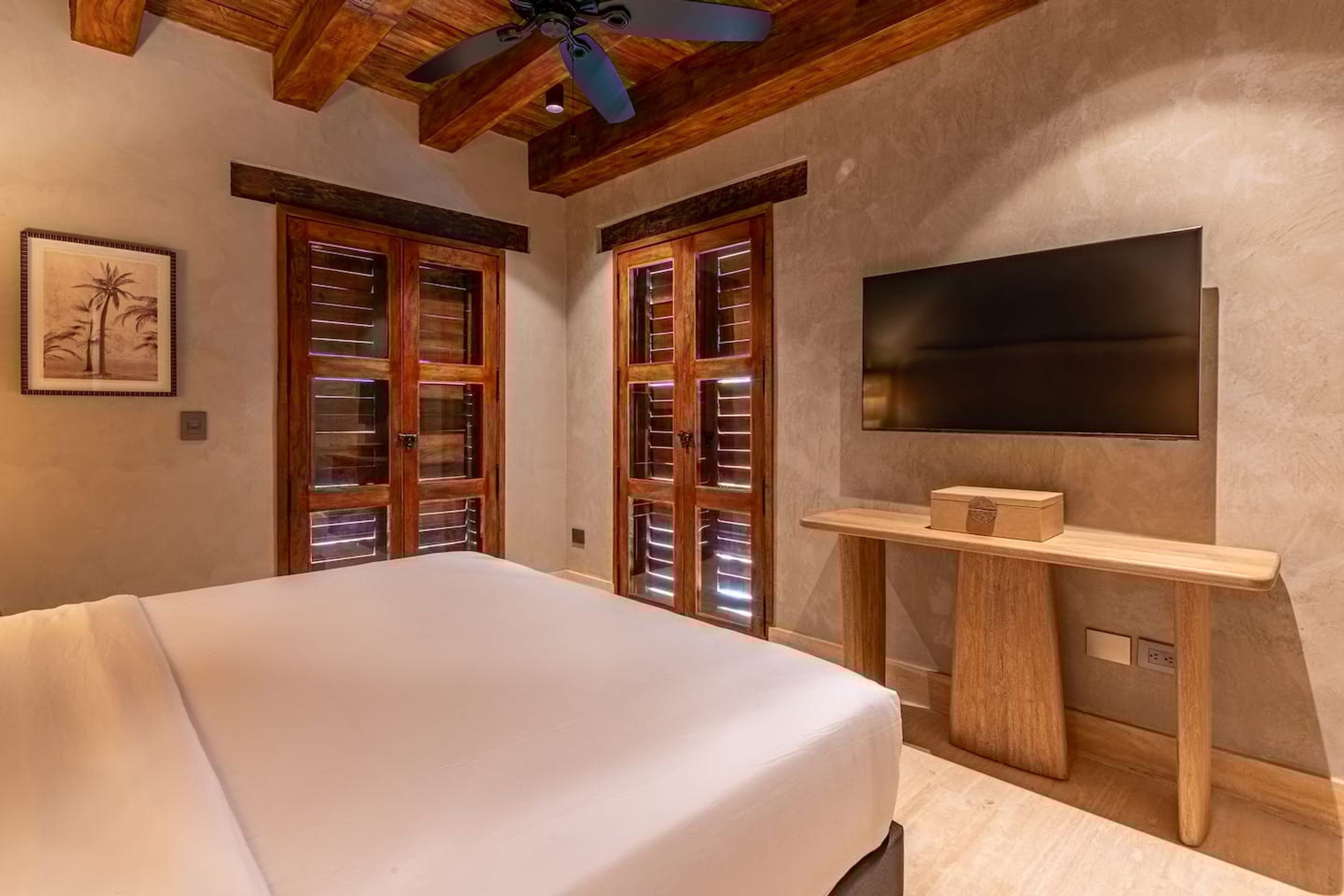 A bedroom with a bed, wooden doors with blinds, a ceiling fan, a TV mounted on the wall, and a small wooden shelf.