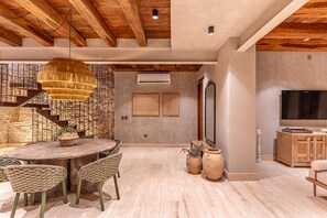 Un salón espacioso con techo de madera, una mesa redonda de comedor con sillas de mimbre, un televisor grande y una escalera con barandilla de hierro forjado.