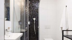A modern bathroom with a glass-enclosed shower, a white sink, and a black shelf with towels. - TurnKey | Angelo Roma Two Level Cozy Studio (Praha 2)
