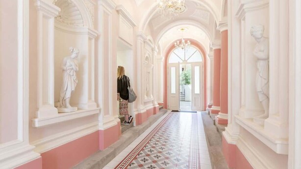 A grand hallway with classical architecture, including columns and statues, leading to a bright doorway. - TurnKey | Angelo Roma Two Level Cozy Studio (Praha 2)