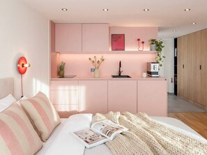 A modern bedroom with a pink theme, including a bed, bedside table, and a small kitchenette area.