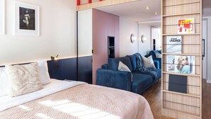 Ein modernes Hotelzimmer mit einem Bett, einem blauen Sofa, Holzboden und einer roten Geländer an einer erhöhten Ebene.