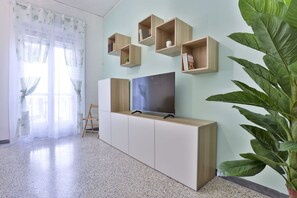 A modern living room with a TV on a low cabinet, wooden shelves on the wall, a large plant, and a window with sheer curtains.