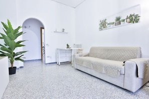 Una habitación con un sofá blanco, una planta en maceta, un radiador y una estantería montada en la pared con plantas.