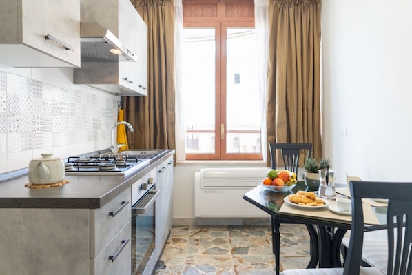 A modern kitchen with a gas stove, a built-in oven, a dining table with a bowl of fruit, and a window with curtains.