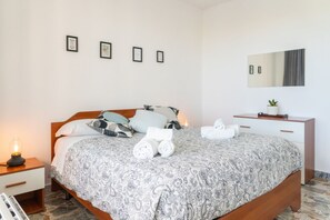 Ein ordentlich bezogenes Bett mit einem gemusterten Überwurf, eine Nachttischlampe, ein kleiner Tisch mit Pflanze und ein Spiegel an der Wand.