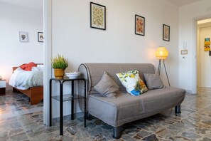 A cozy room with a grey sofa, a small side table with a plant, a lamp, and a bed with a red blanket.