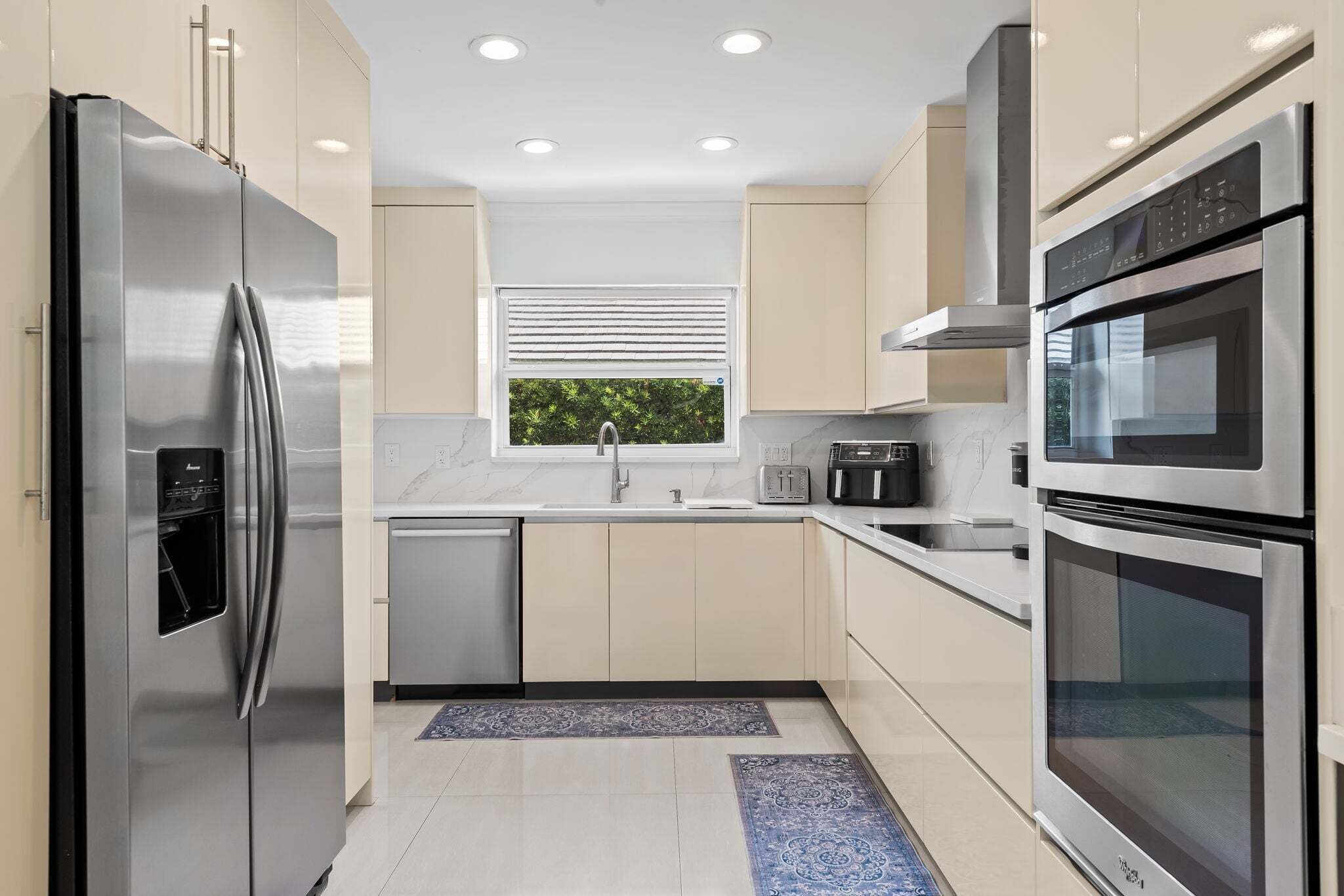 A modern kitchen with stainless steel appliances, a window with blinds, and a view of greenery outside.