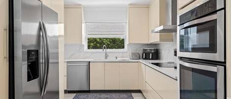 A modern kitchen with stainless steel appliances, a window with blinds, and a view of greenery outside.