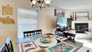 A dining area with a wooden table, four chairs, and a chandelier. There are two windows with blinds, a fireplace, and wall decorations including maps and a painting.