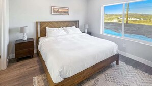 A bedroom with a large bed, a nightstand, a lamp, a framed picture on the wall, and a window with a view.