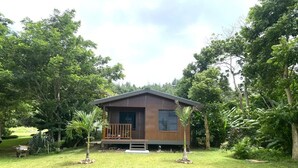 Ein kleines, eingeschossiges Holzhaus mit Veranda, umgeben von üppigem Grün und Bäumen.