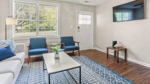 Een woonkamer met een witte bank, blauwe stoelen, een salontafel, een televisie en een plafondventilator.