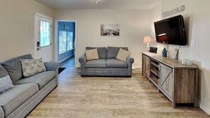 A living room with a grey sofa, a grey couch, a wooden cabinet, and a flat-screen TV on the wall. - The Charming Cozy Cottage by Miss Kitty’s (Rockport)