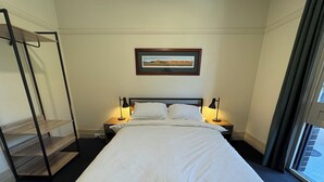 A neatly made bed with white linens, two bedside lamps, a framed picture on the wall, and a window with curtains. - The Bat & Ball Hostel (Redfern)