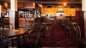 A restaurant interior with a bar area, multiple tables and chairs, and a patterned carpet. - The Bat & Ball Hostel (Redfern)