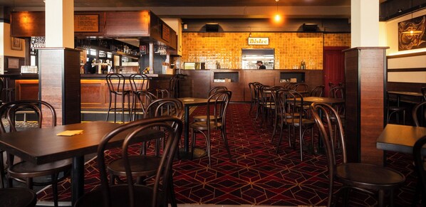 A restaurant interior with a bar area, multiple tables and chairs, and a patterned carpet. - The Bat & Ball Hostel (Redfern)
