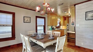 Zona pranzo con tavolo in legno e sedie bianche, un lampadario e una cucina sullo sfondo.