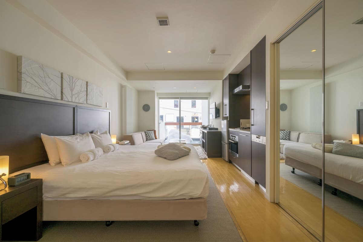 A spacious hotel room with a large bed, a bedside table with a lamp, a wardrobe, and a view through a glass door.