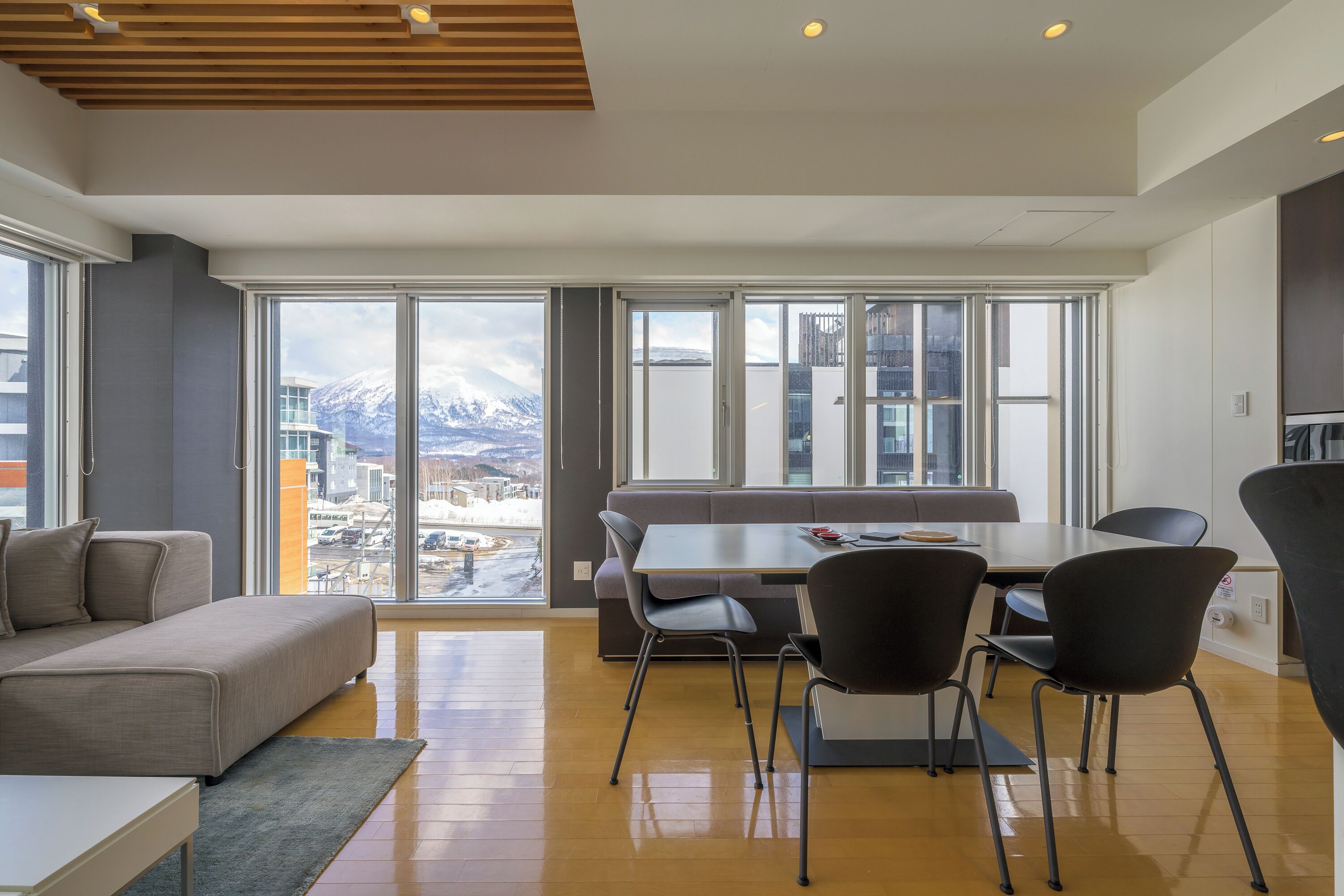 Sala de estar moderna com sofá, mesa de jantar e cadeiras, grandes janelas com vista para a cidade e montanhas, e teto de madeira.