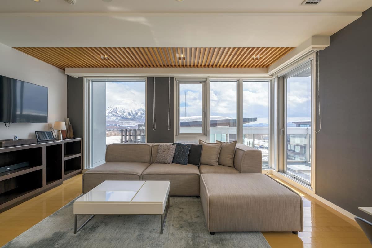 A modern living room with a sectional sofa, a coffee table, and a flat-screen TV. Large windows offer a view of a snowy mountain landscape.