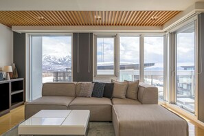 A modern living room with a large sectional sofa, a coffee table, and a view of snow-capped mountains through large windows. - Alpen Ridge (Kutchan)