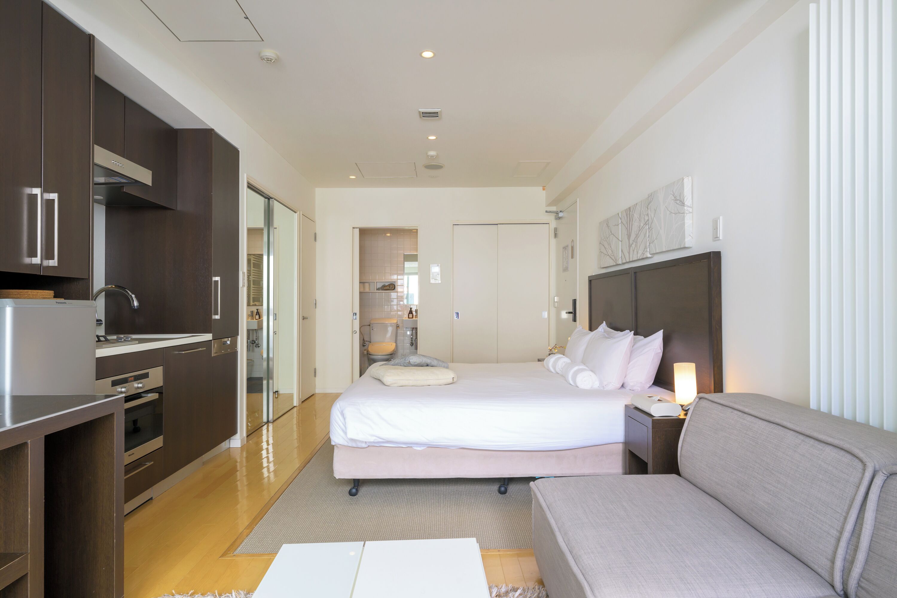 A modern hotel room with a bed, a sofa, a desk, and a kitchenette.