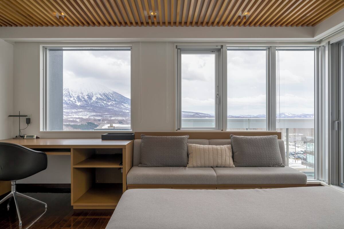 A modern bedroom with a large bed, a desk, and a sofa. There are large windows offering a view of mountains and a cityscape.