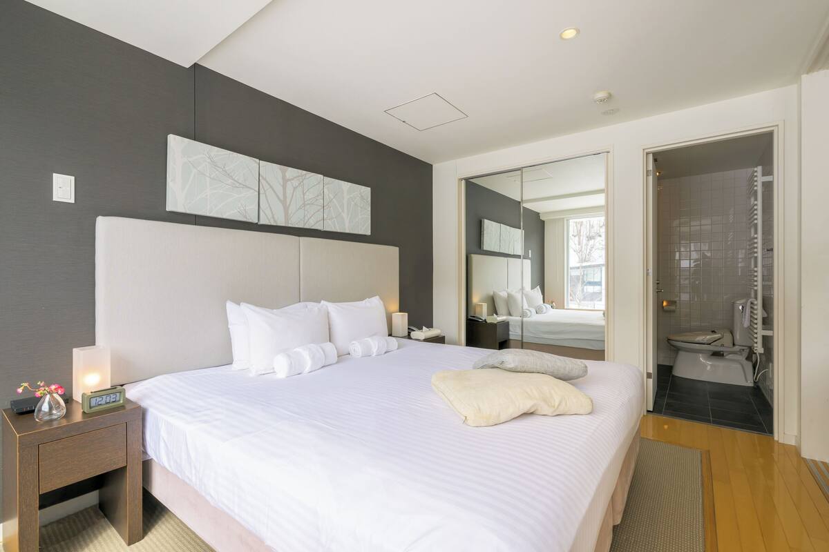 A modern hotel room with a large bed, a nightstand with a lamp, a mirror, and a view into the bathroom.