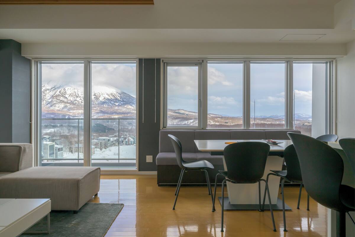 A modern living room with a large window, a sofa, a dining table with chairs, and a view of snow-capped mountains.