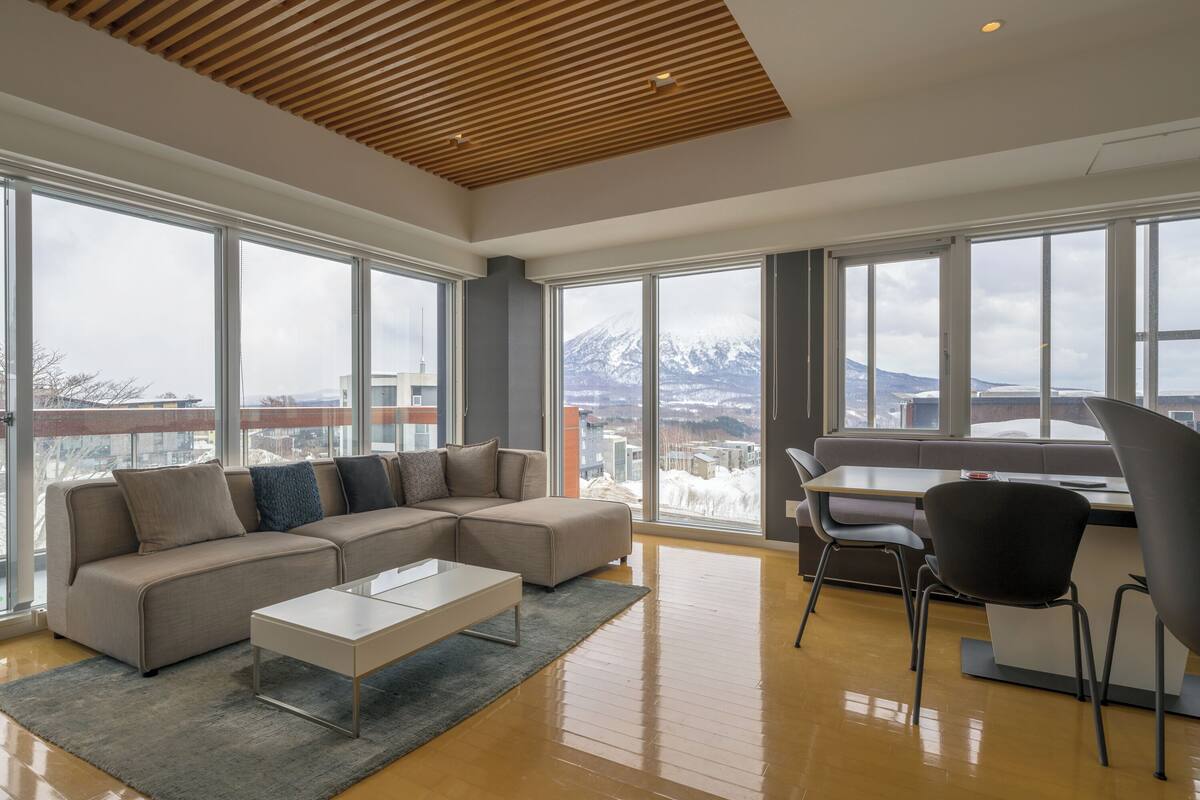 A modern living room with a sectional sofa, a coffee table, and a dining area with chairs. Large windows offer a view of a snowy mountain and cityscape.