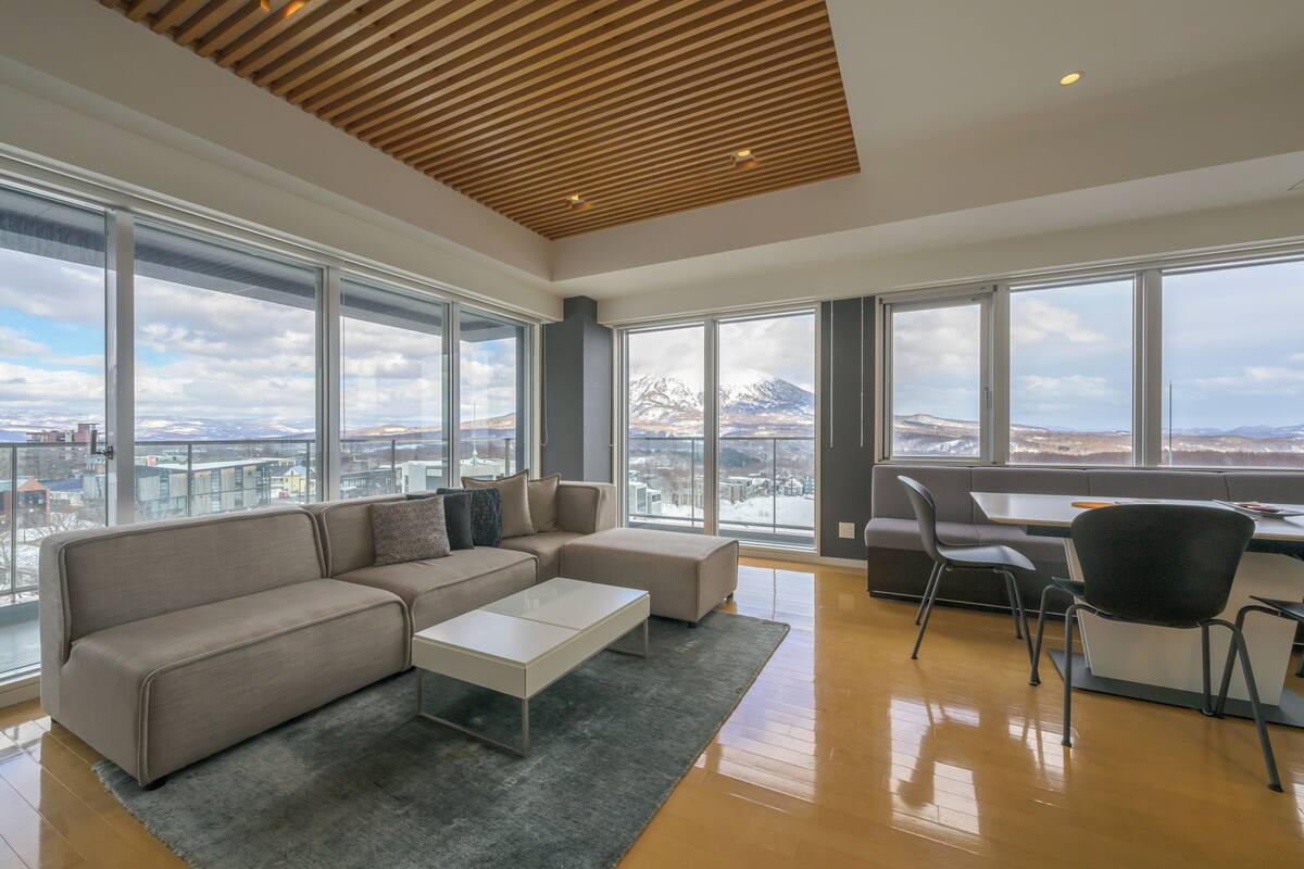 A spacious living area with a large sectional sofa, a coffee table, and a desk with chairs. Large windows offer a view of a snowy mountain range.