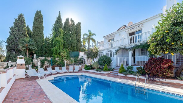 Une piscine dotée d’une terrasse carrelée, entourée d’une végétation luxuriante et bordée d’un bâtiment blanc avec un balcon.