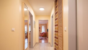Un couloir avec un sol carrelé, une porte en bois ornée d’un rideau à carreaux, et une pièce visible au fond, avec un canapé et une fenêtre.