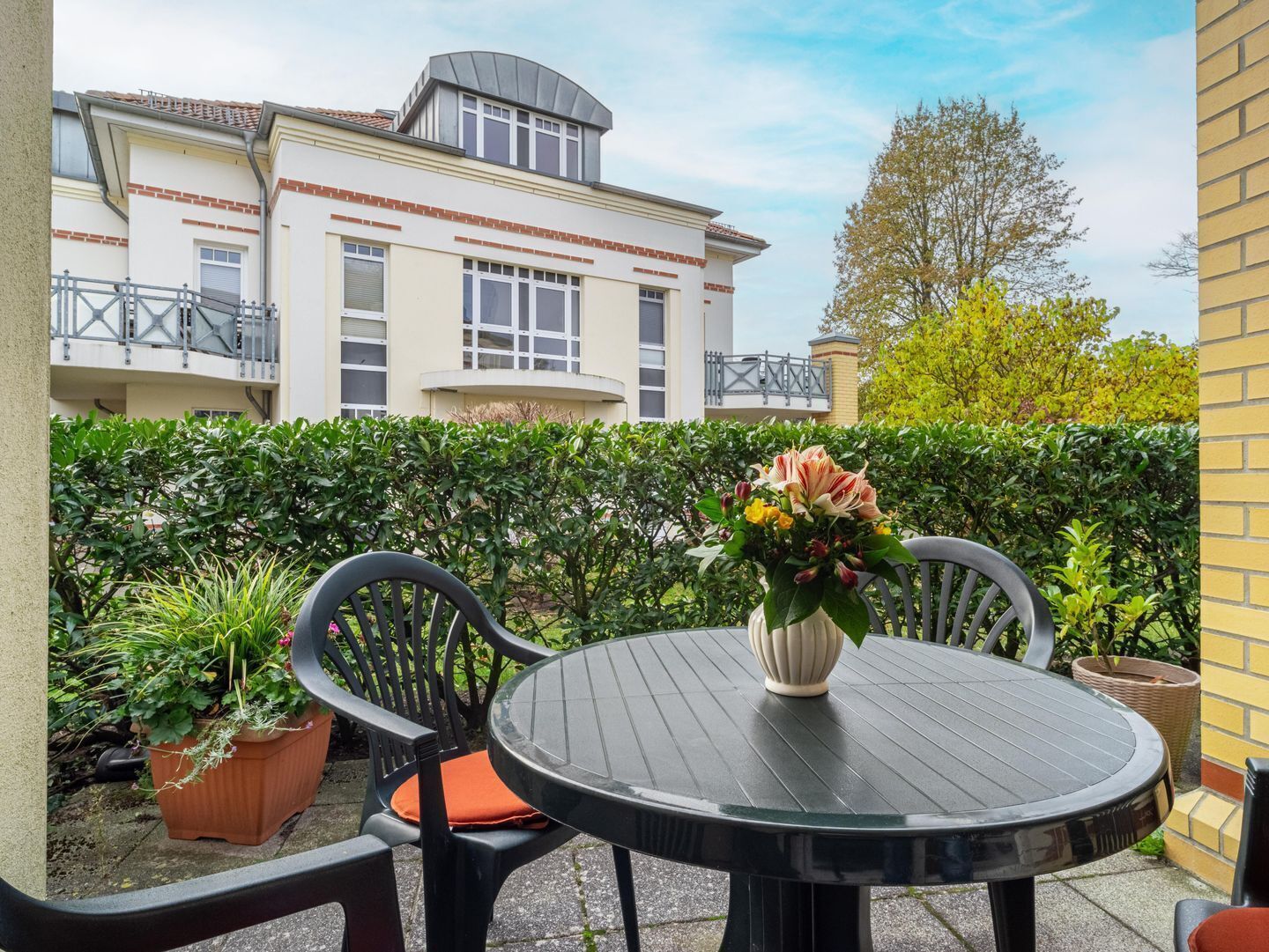 A patio with a round table and chairs, a potted plant, and a building in the background.