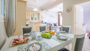 A dining area with a table set for a meal, chairs, a kitchen in the background, and a living area with a sofa and a clock on the wall.