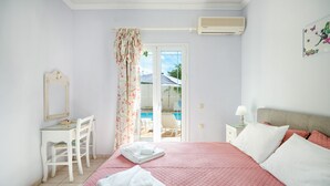A bedroom with a bed, a desk, a chair, a nightstand, and a window with a view of a pool.