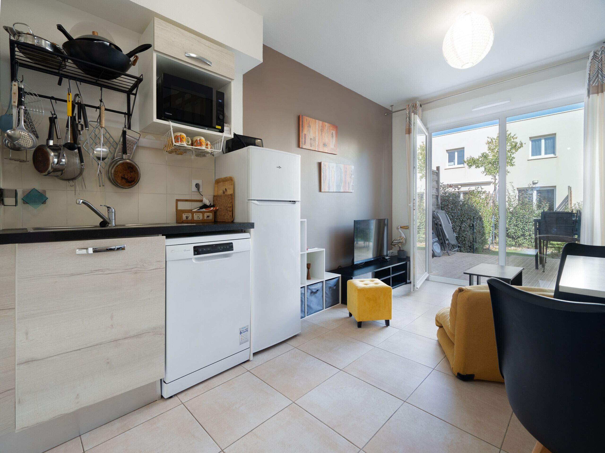 Een moderne keuken met een vaatwasser, magnetron en diverse keukengerei. Er is een eethoek met een gele poef en een televisie.